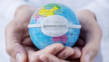Fotografia de um homem segurando um pequeno globo terrestre na palma das mãos. O globo aparece usando uma máscara de proteção onde se lê coronavírus